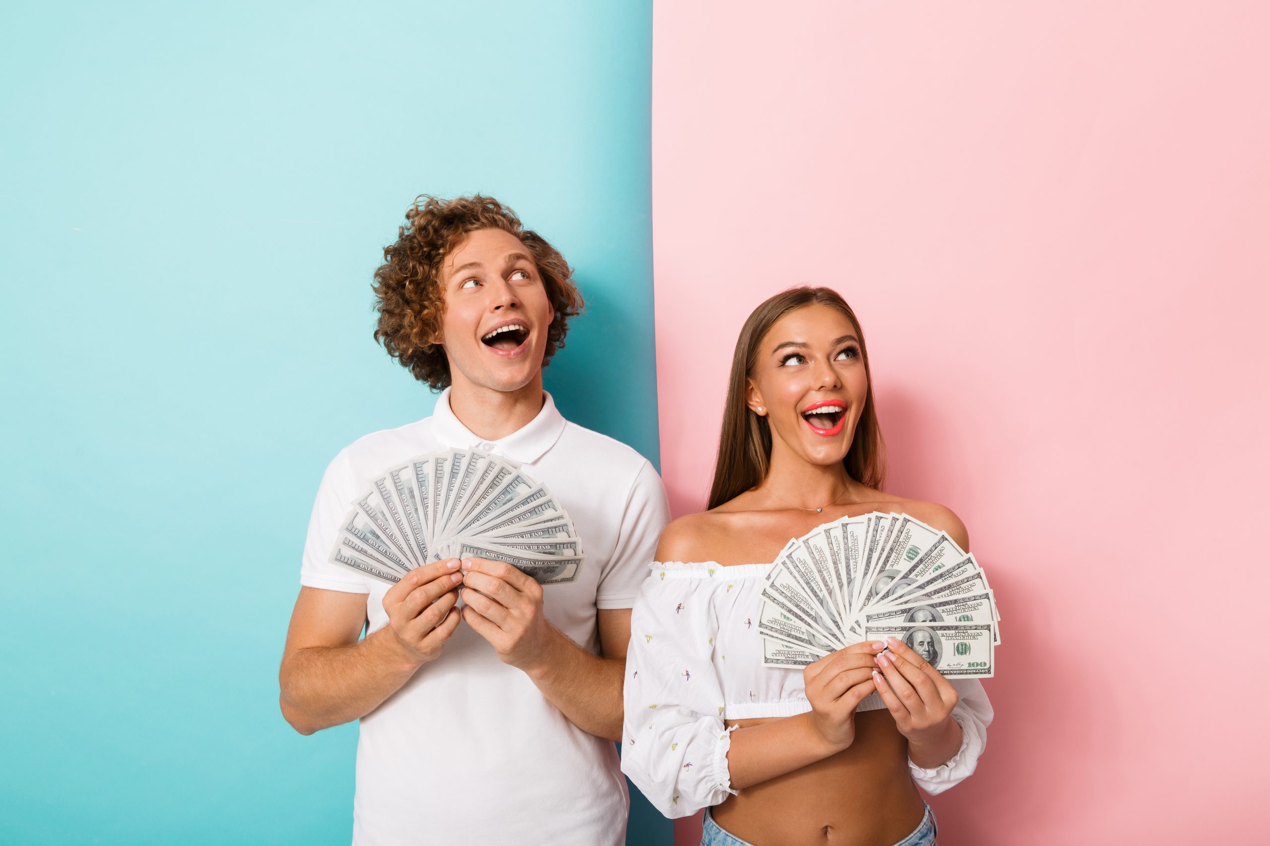 Portrait of a cheerful young couple standing over two colored background, showing money banknotes, looking up