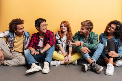 Multiethnic group of smiling young people sitting on the floor and talking over yellow background