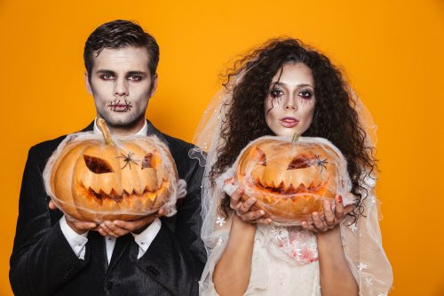 Photo of scary zombie couple bridegroom and bride wearing outfit and halloween makeup holding carved pumpkin isolated over yellow background